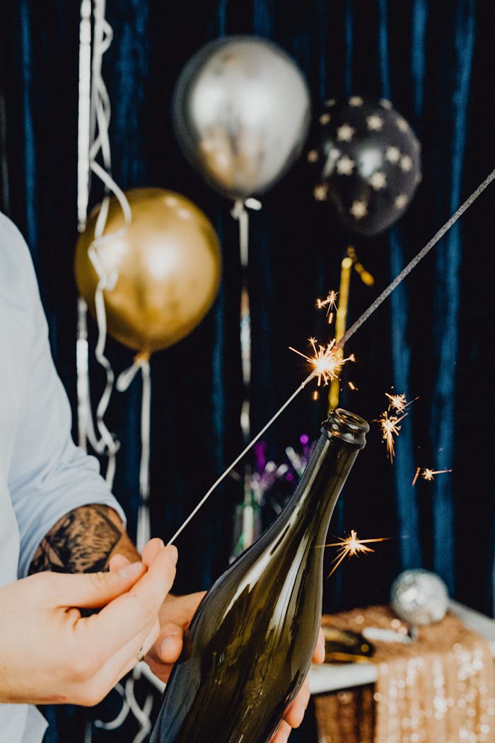 Tattooed hands holding a sparking sparkler near a champagne bottle at a festive party.